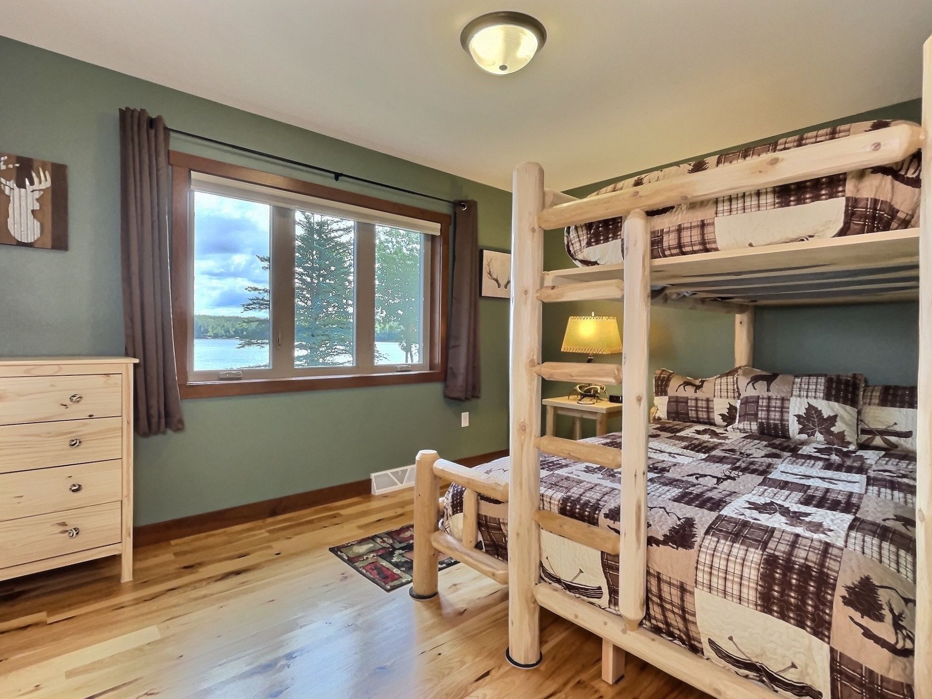 Bedroom with bunk beds, dresser, and window overlooking a lake. Green walls, wooden floors, and lodge-style bedding.