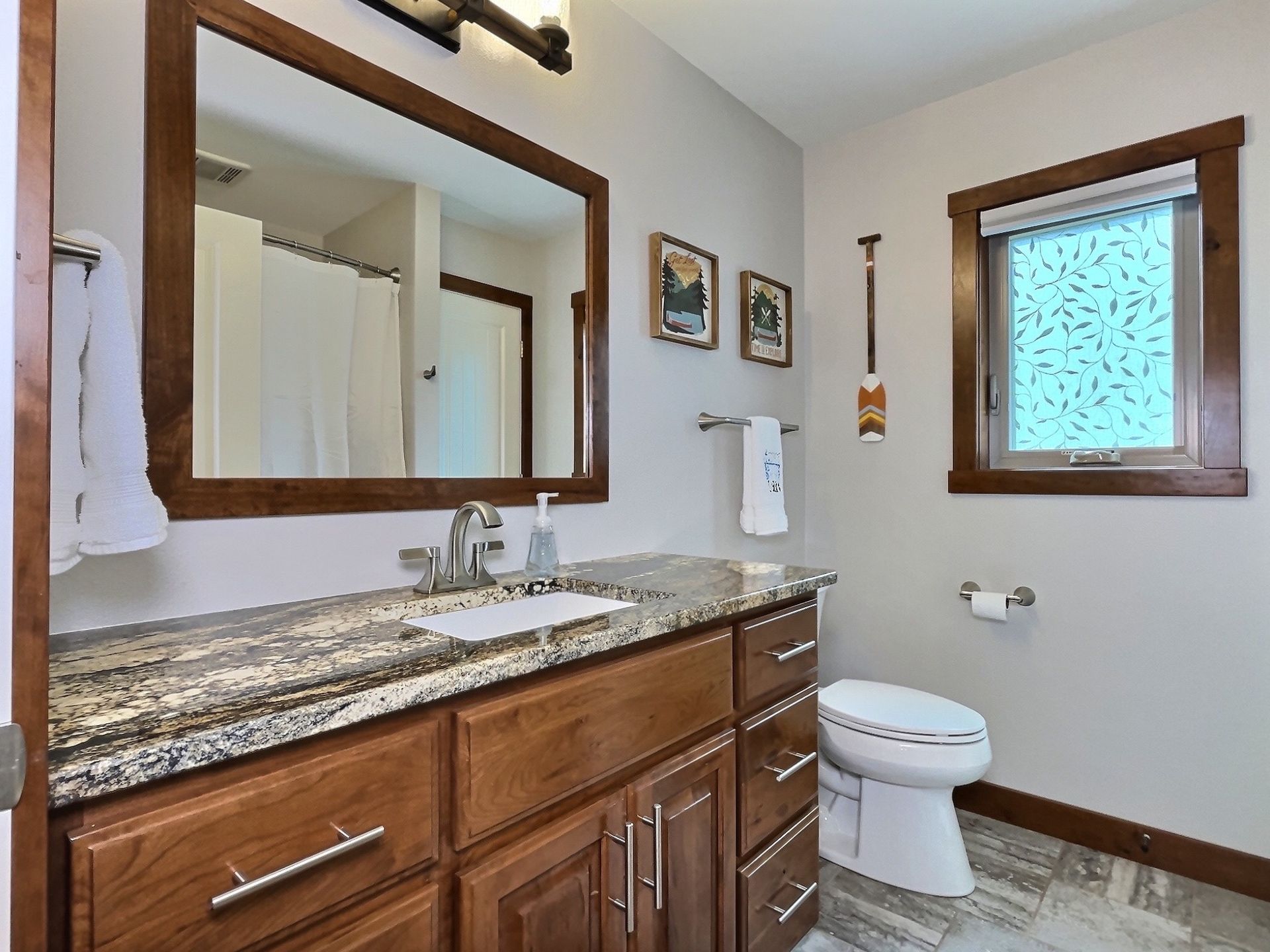 Bathroom with brown vanity, mirror, toilet, and a window with a patterned shade; light gray walls.