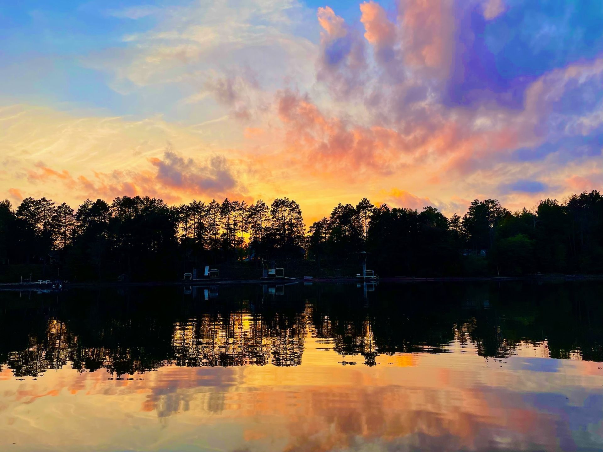 Sunset over calm water reflects colorful clouds and silhouetted trees.