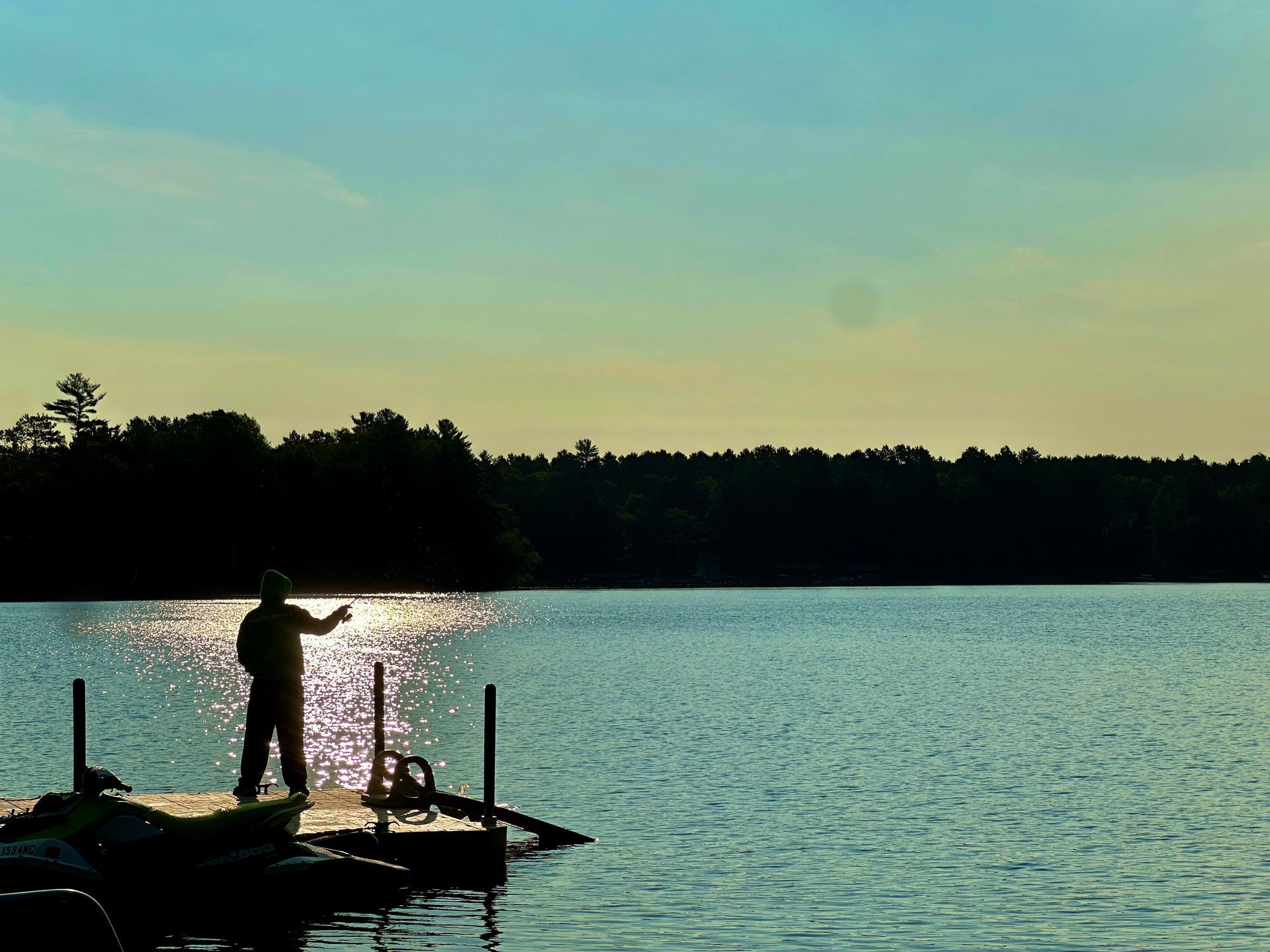 Person fishing on a dock silhouetted against a shimmering lake at sunset.