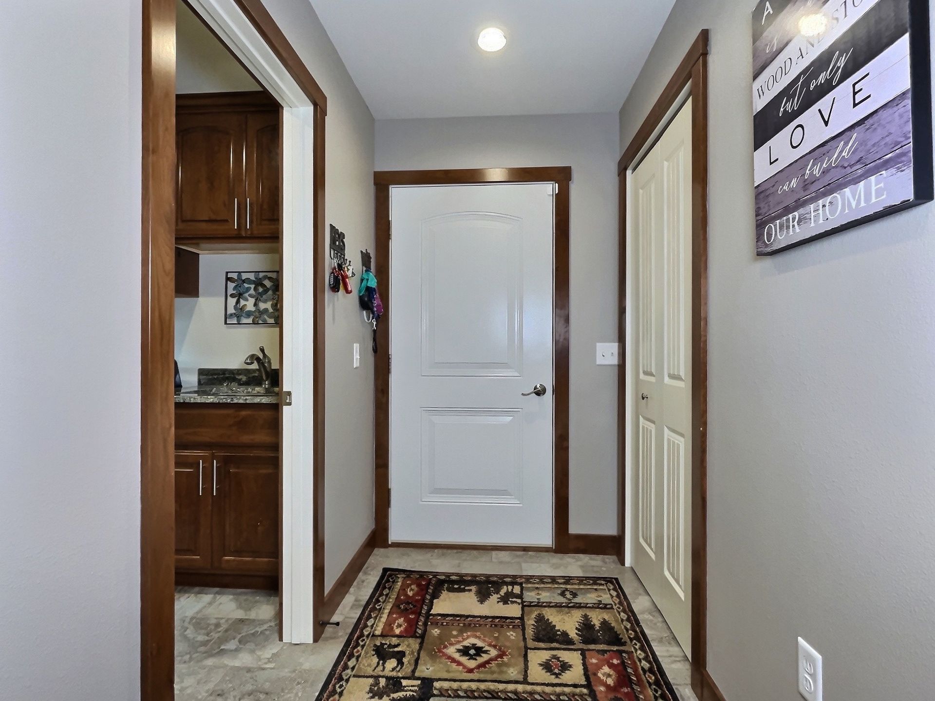 Hallway with white door, rug, and artwork. A doorway leads to a kitchen. Wooden trim frames the walls and doors.