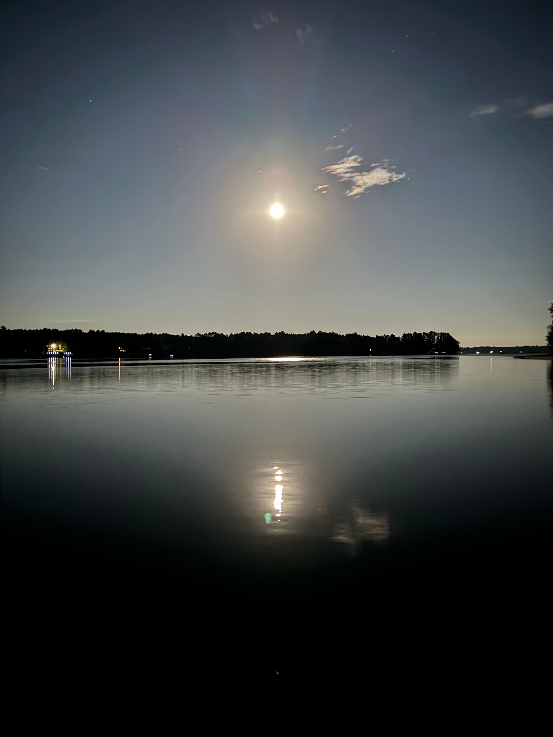Sun reflecting on a calm lake, with dark shoreline and blue sky.