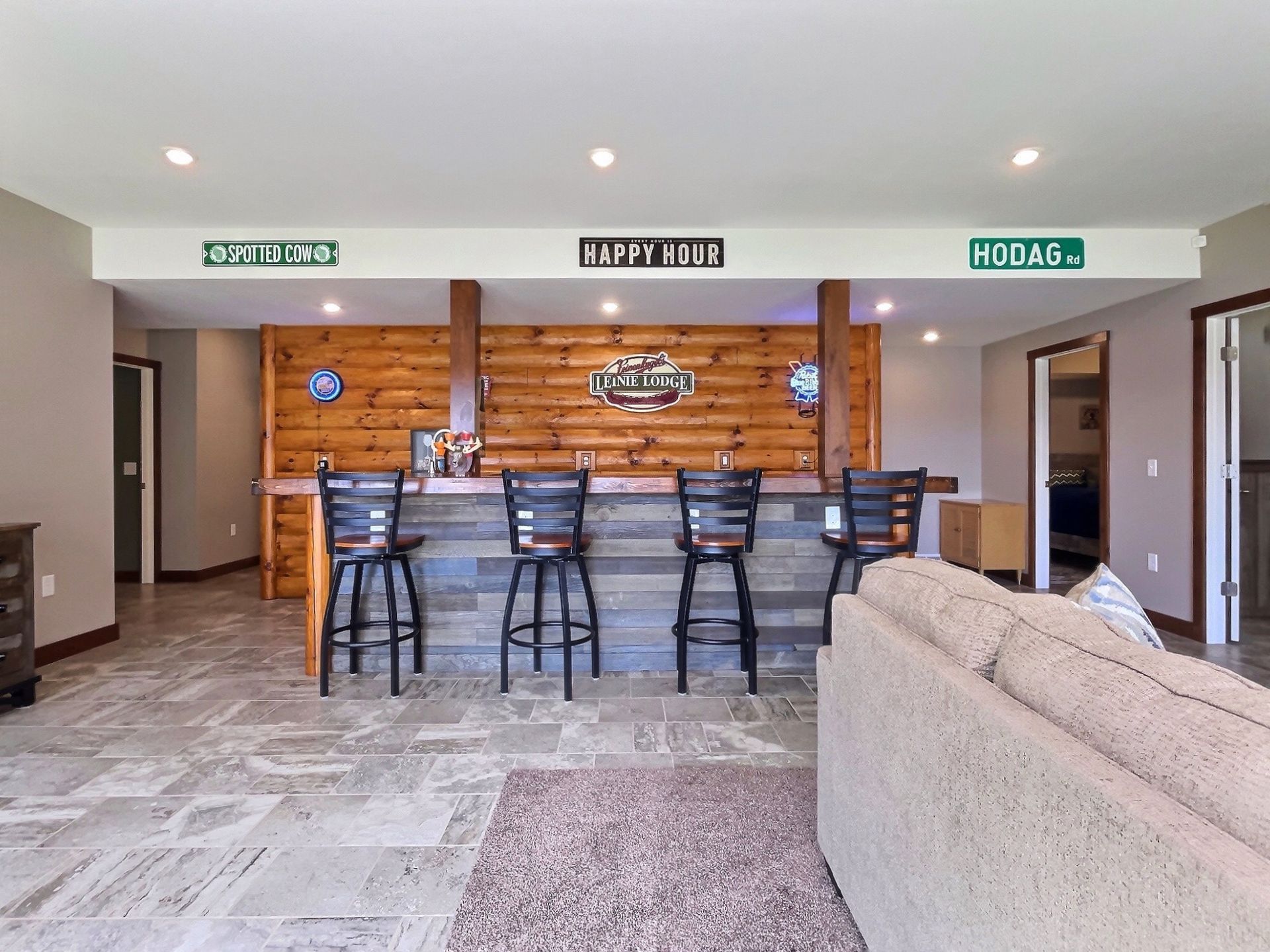 Bar with wooden facade, stools, and signs, lit by recessed lights. Stone floor with a rug and a couch.
