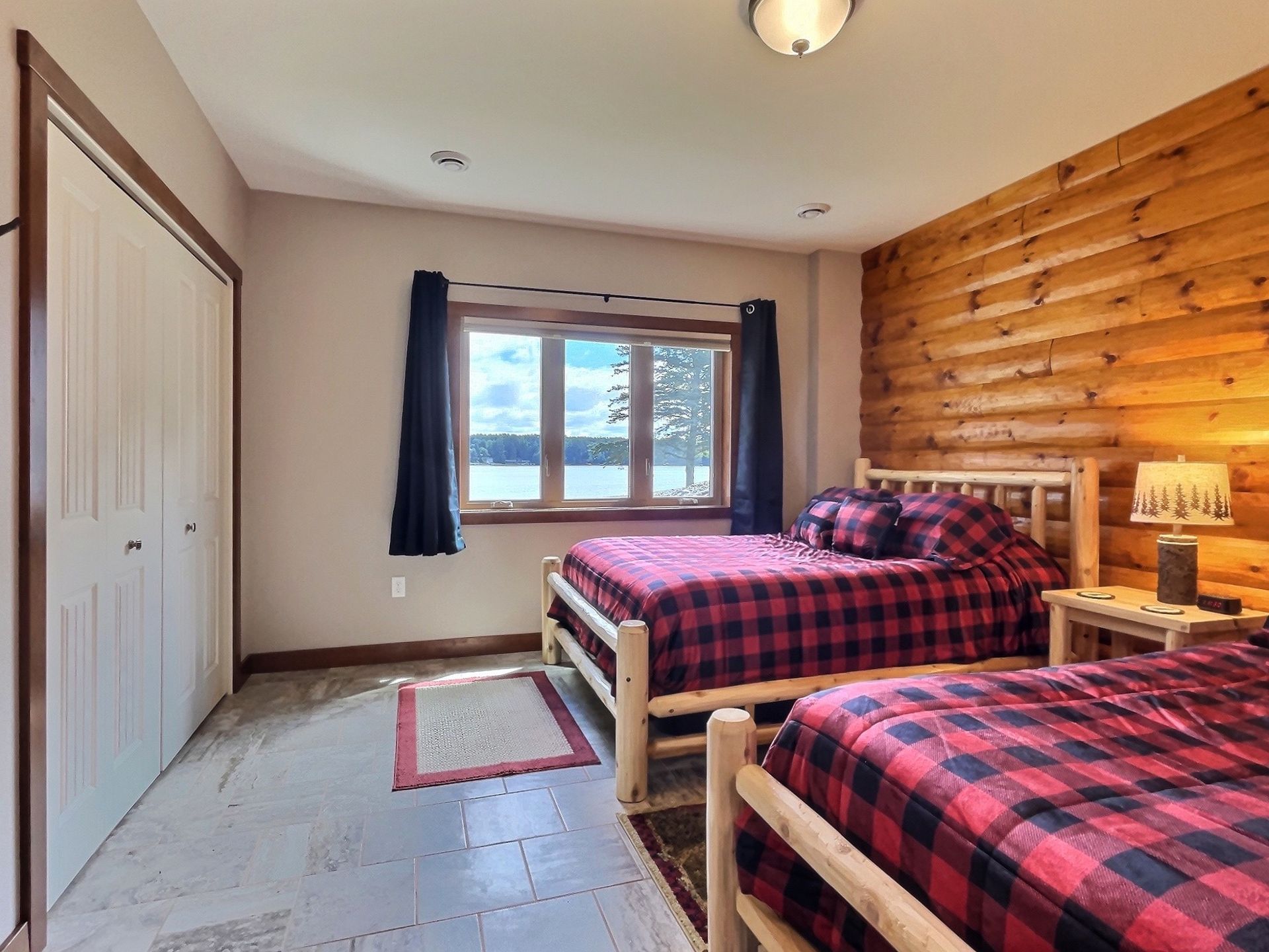 Bedroom with two beds, red plaid bedding, log accent wall, and a window with a lake view.