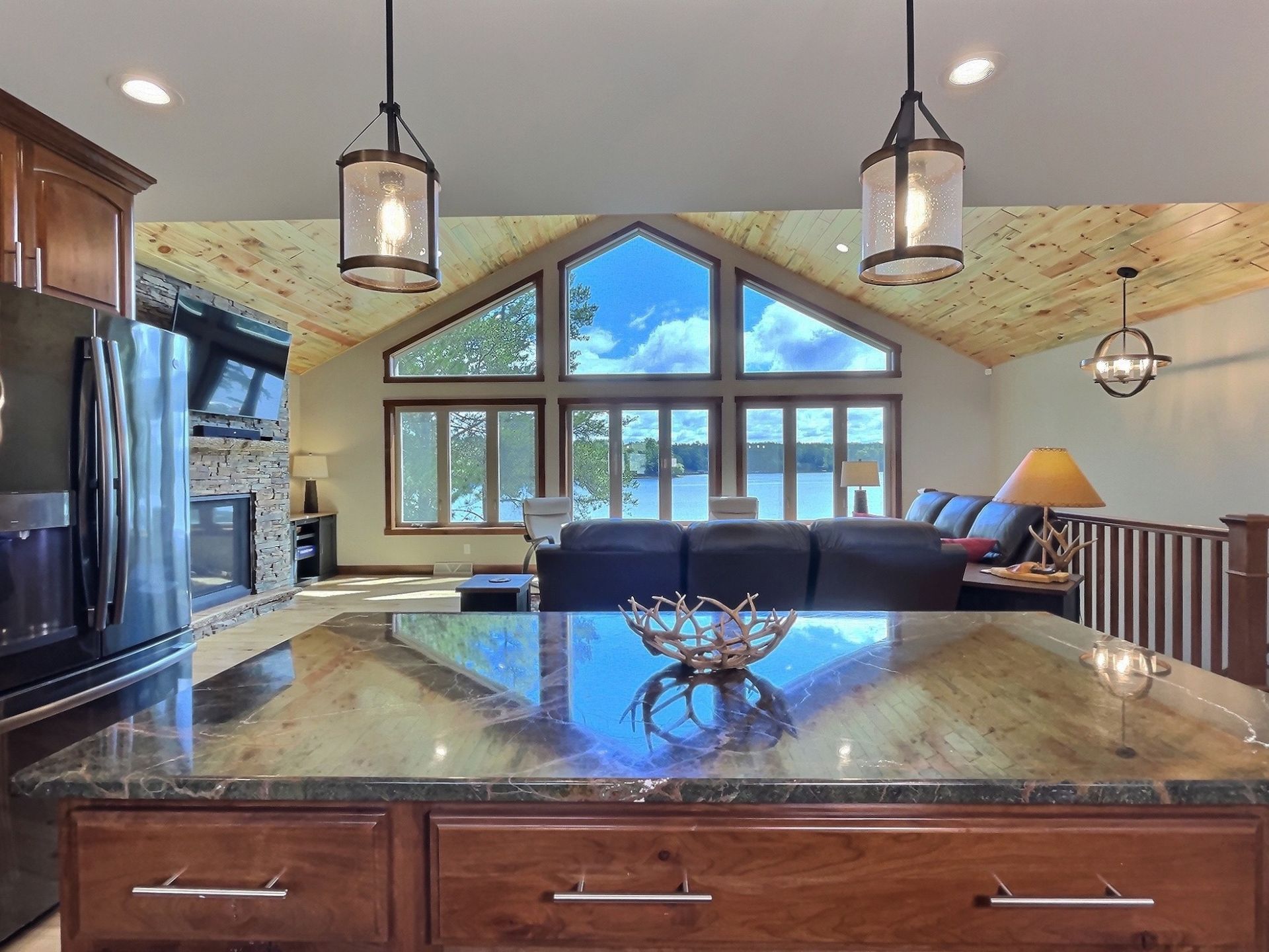 Interior with kitchen island and view of a lake through large windows. Pendant lights, fireplace, and sofa visible.