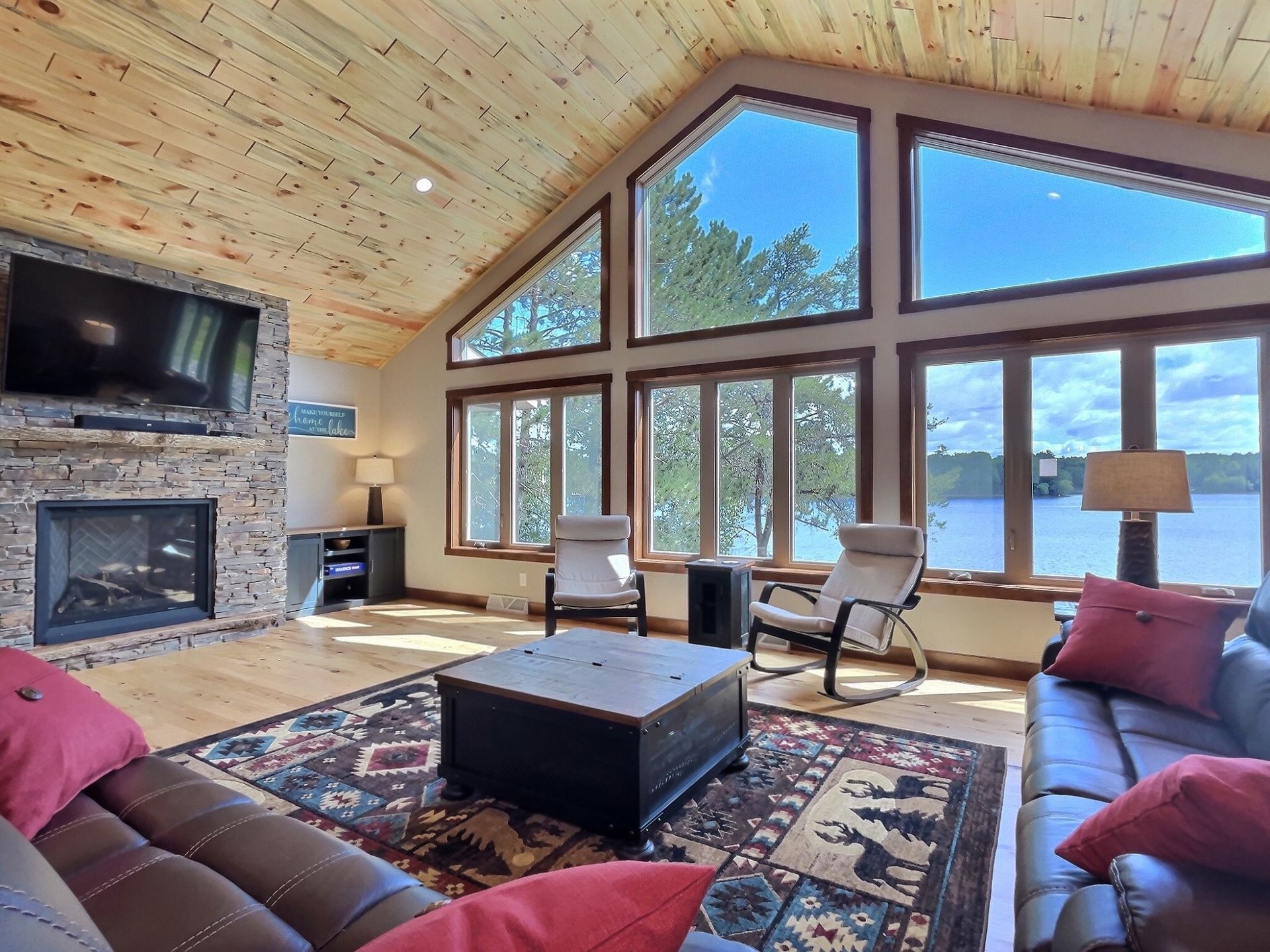 Living room with lake view, fireplace, leather sofas, chairs, wooden ceiling and floor, and large windows.