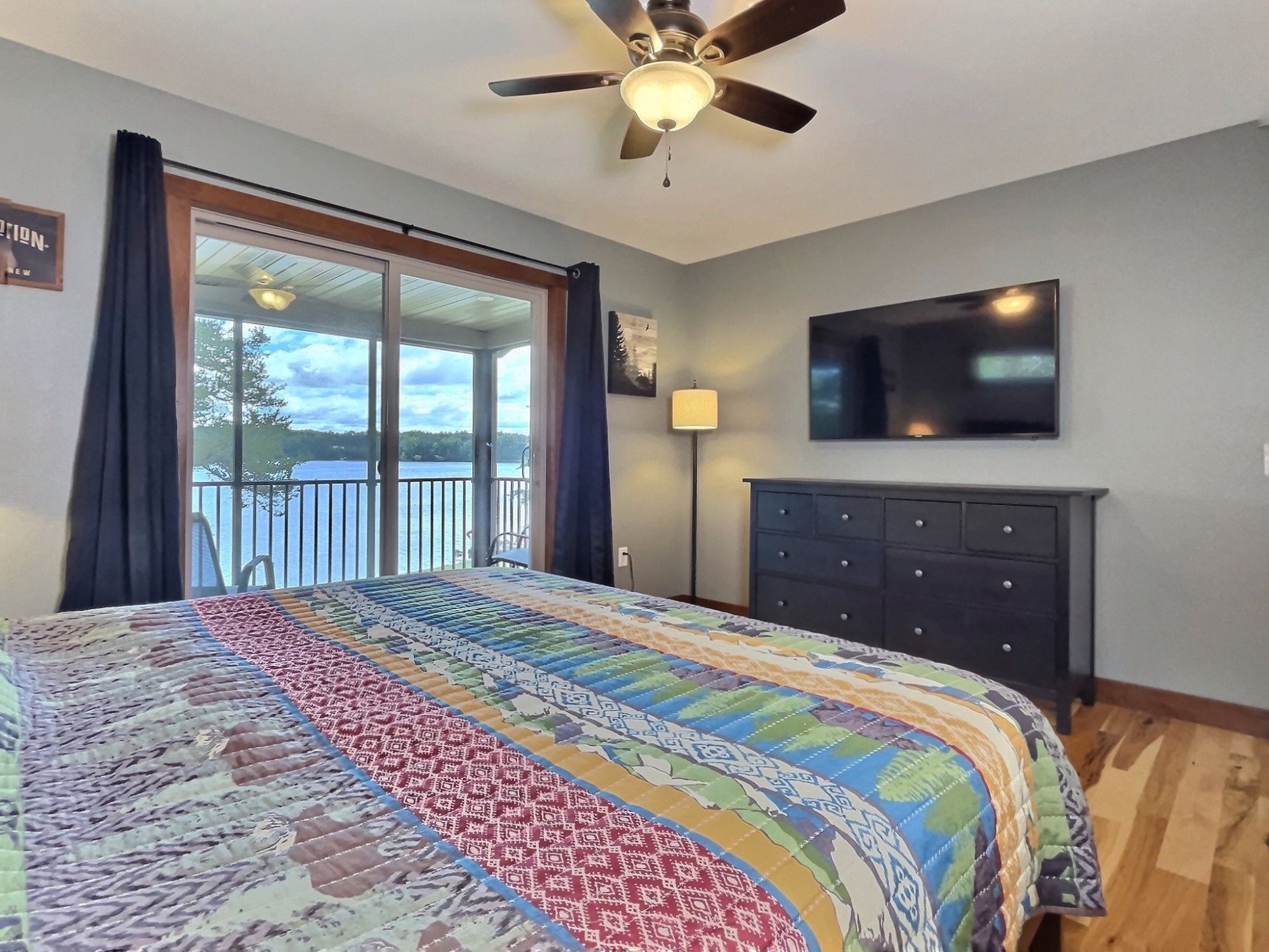 Bedroom with a bed, TV, dresser, and a sliding glass door leading to a balcony overlooking water.