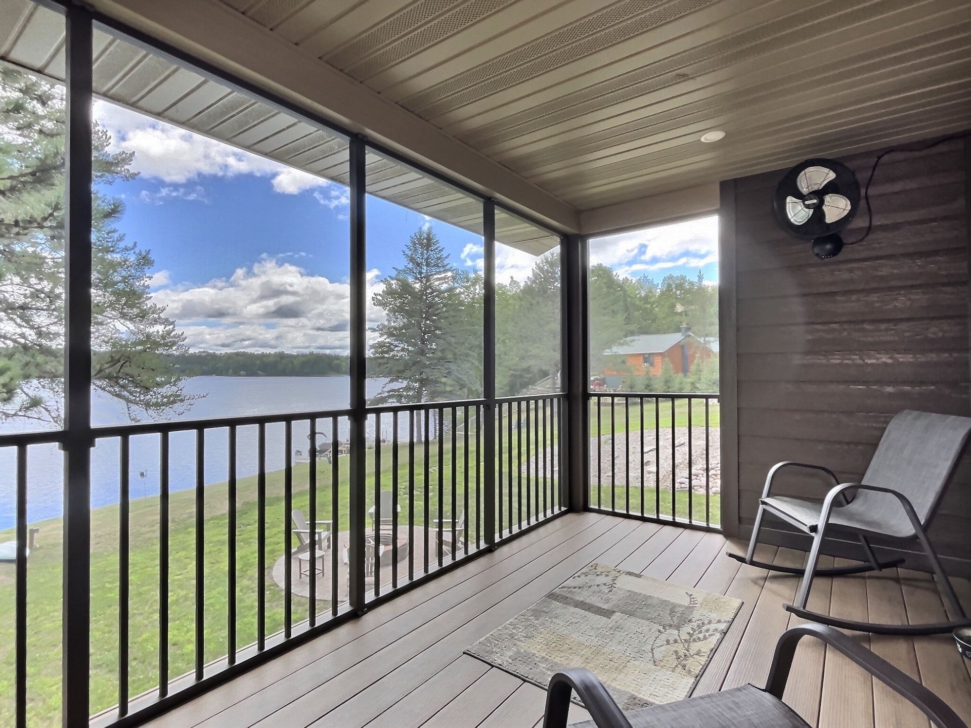 Screened porch with lake view, black railing and frames, two rocking chairs, fan, blue sky.