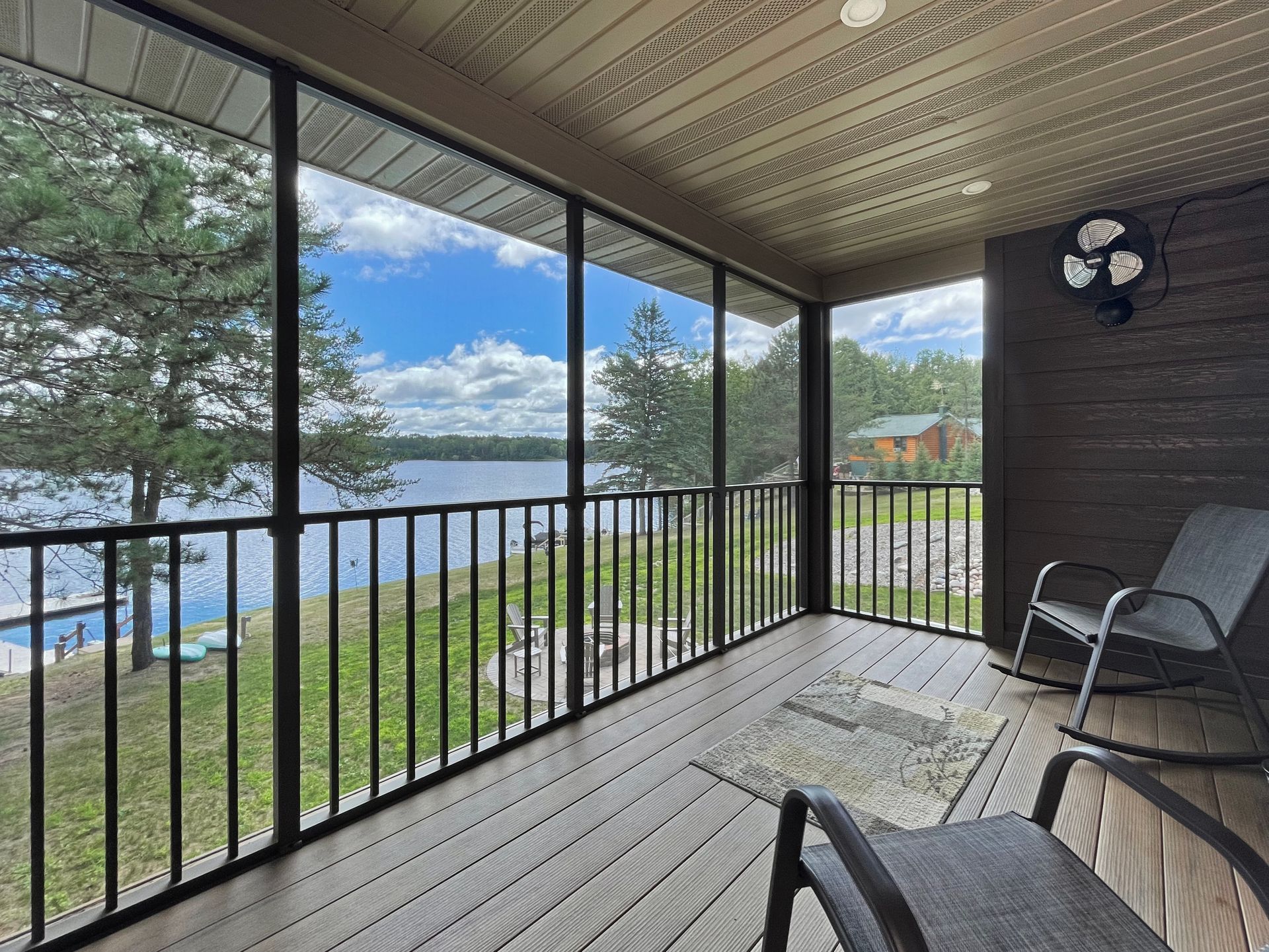 Screened porch overlooking a lake with two rocking chairs. Blue sky, water, and trees visible.