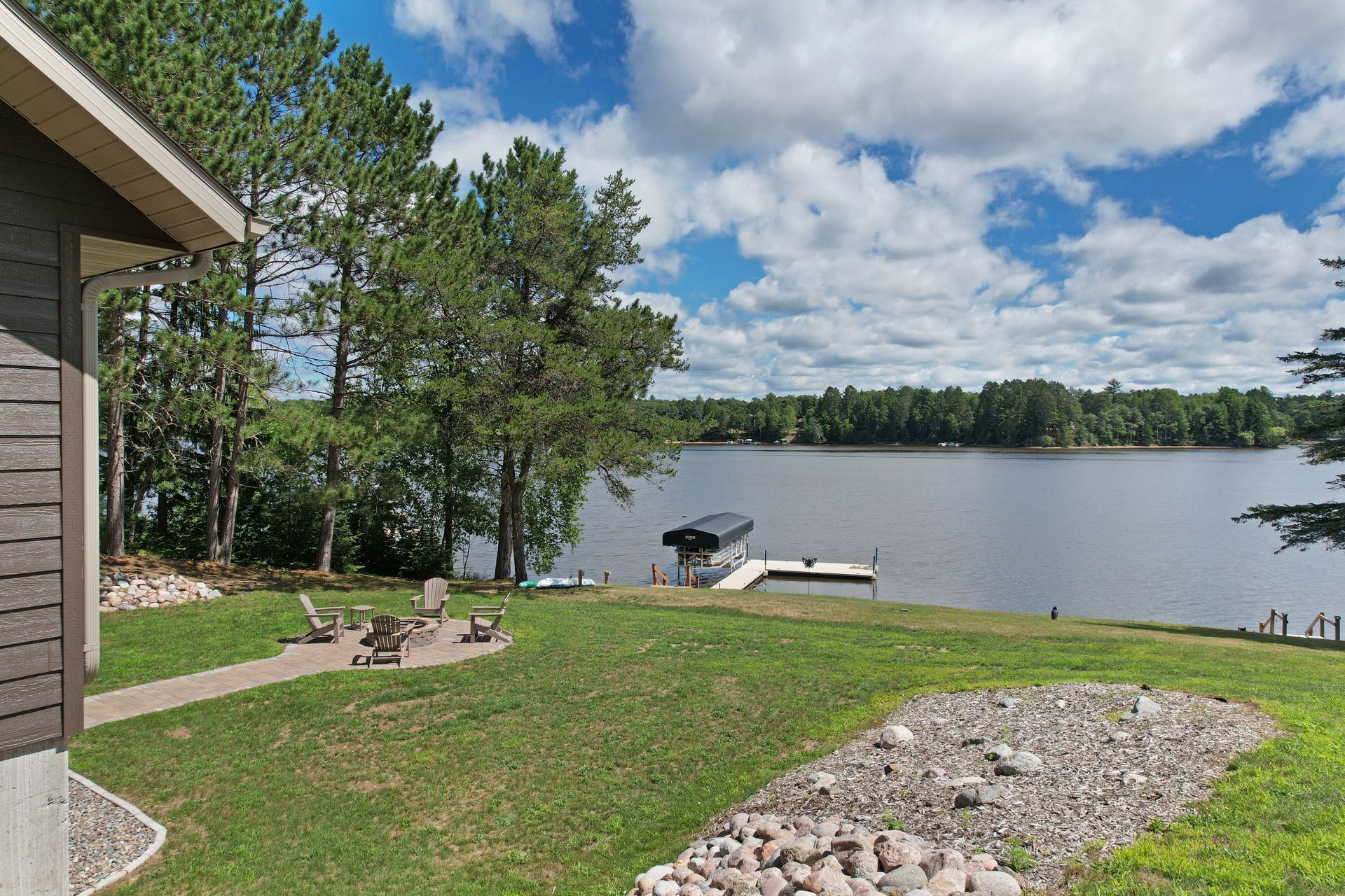 Lakeside view with green grass, trees, and dock. House on left, blue sky with clouds.