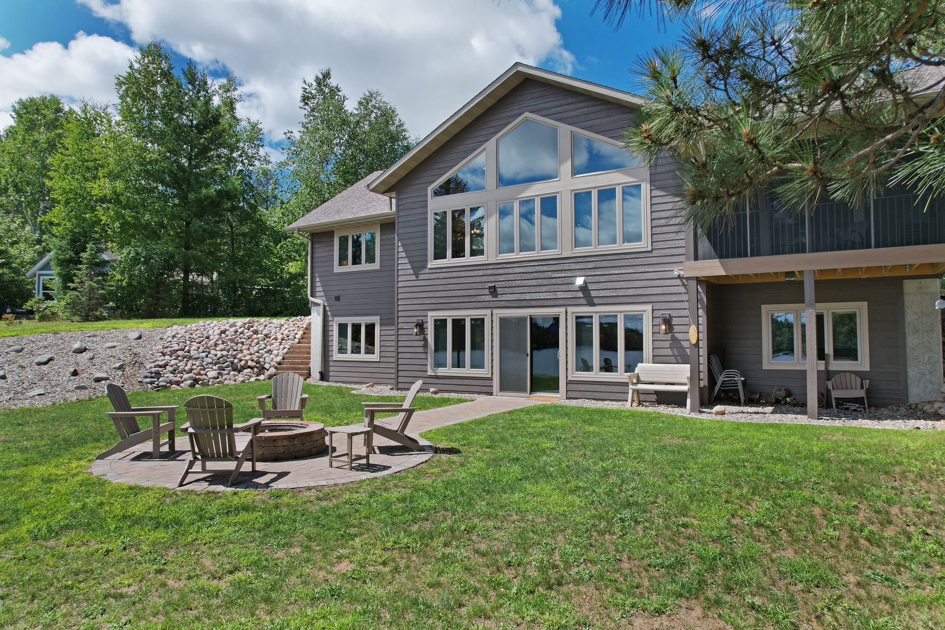 Gray house with large windows, fire pit, and lawn overlooking a lake on a sunny day.