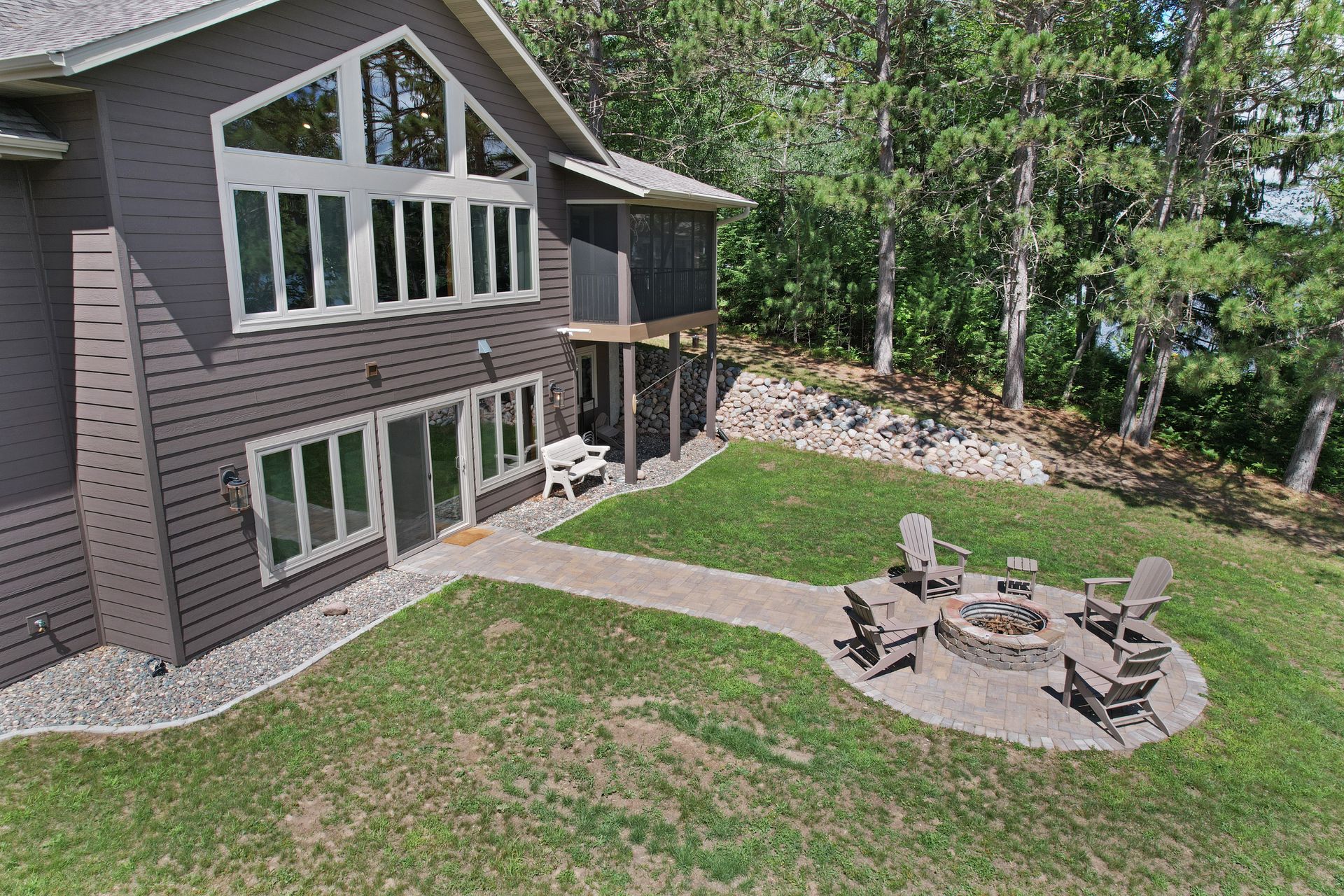 House with large windows, screened porch, fire pit, and Adirondack chairs on a grassy hill overlooking a lake.