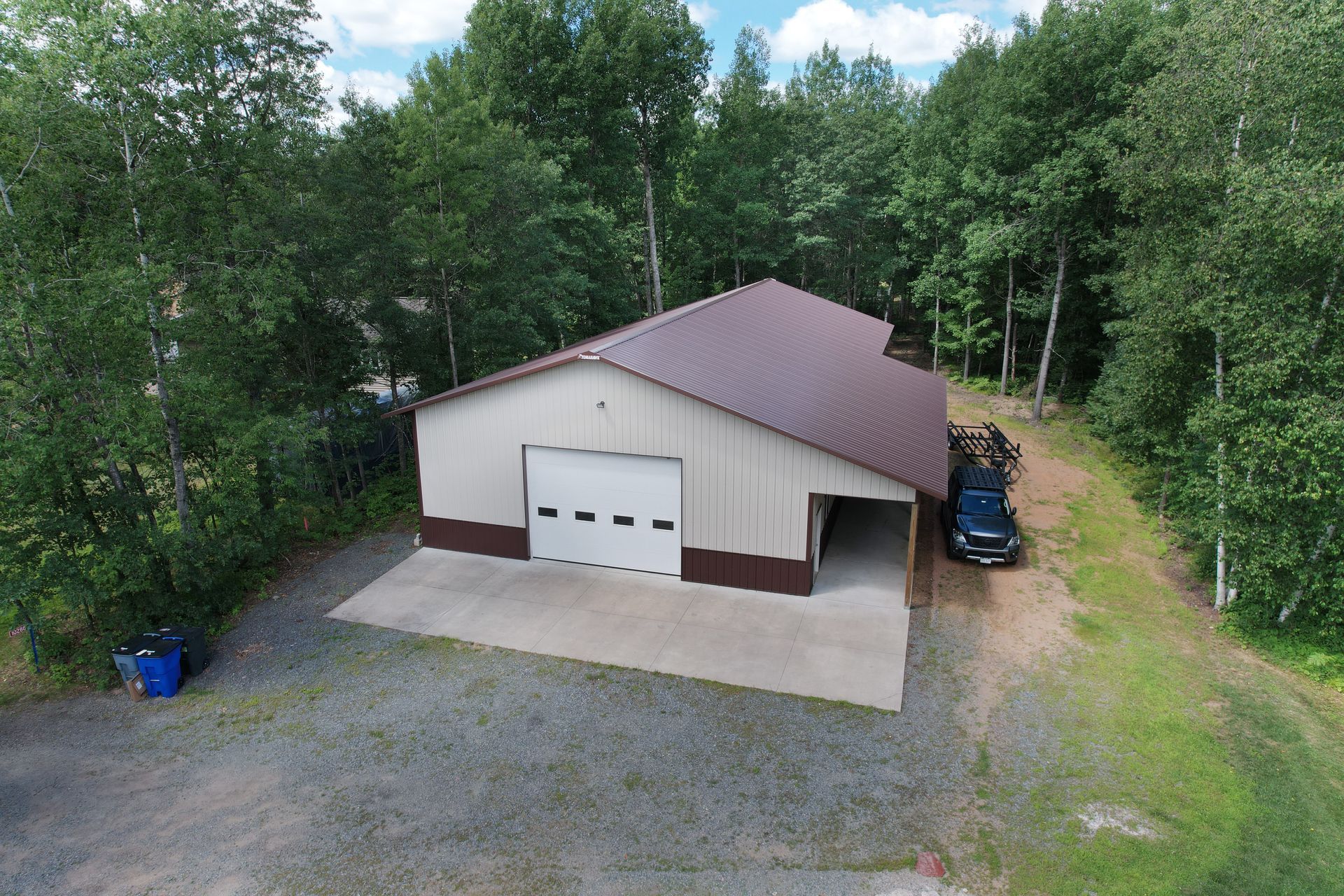 Steel-frame building with brown roof and trim, white garage door, open car bay; car parked nearby, surrounded by trees.