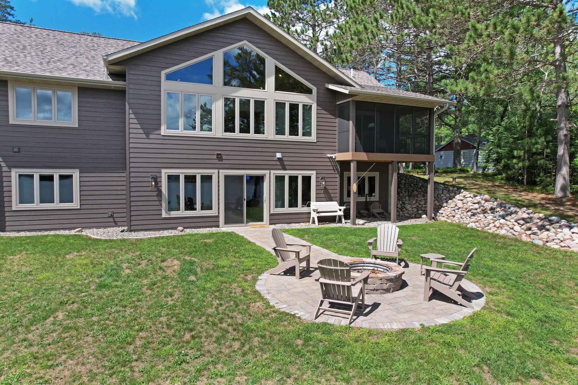 Brown house with large windows, a screened porch, and a patio with a fire pit, overlooking a grassy yard.