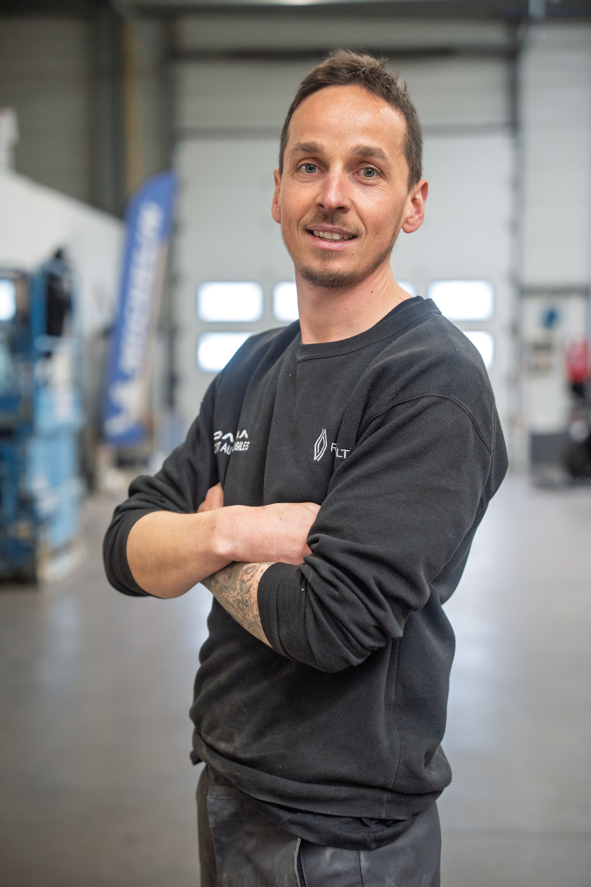 Jeune homme dans un atelier croisant les bras et portant un pull noir avec le logo Renault