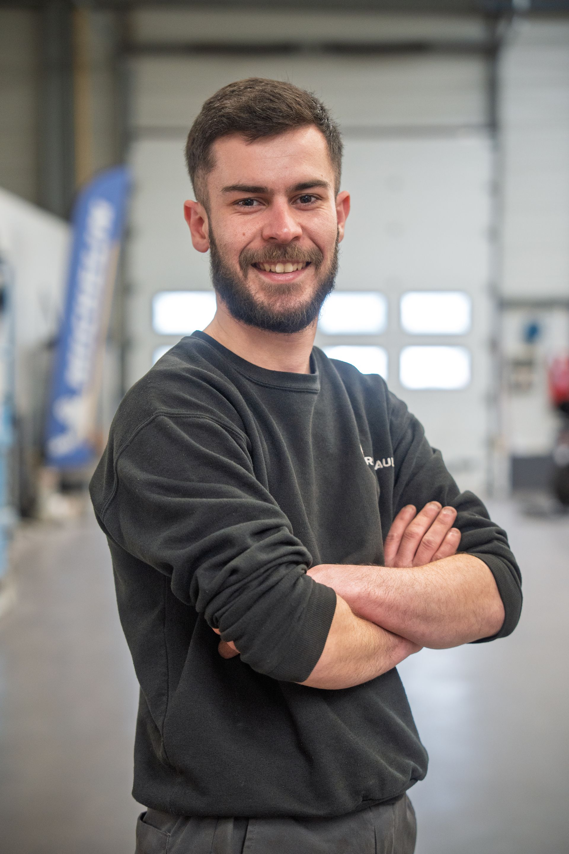 Jeune homme barbu et souriant dans un atelier croisant les bras et portant un pull noir à manches longues