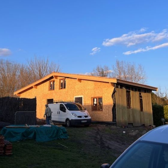Maison en bois beige de deux étages en construction, avec une camionnette blanche garée devant sous un ciel bleu.