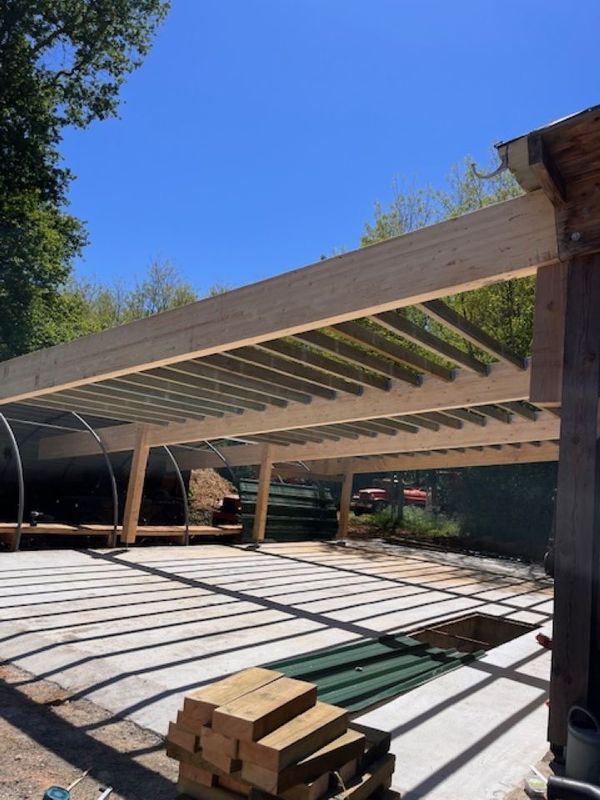 Pergola en bois partiellement construite dans une cour ensoleillée, avec du bois empilé au premier plan.