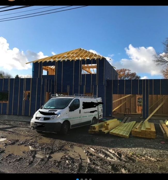 Une camionnette blanche est garée à côté d'une maison bleue en construction, dont la charpente du toit est apparente et des planches jonchent le sol.