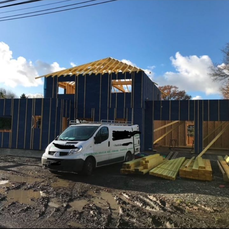 Une camionnette blanche est garée devant une maison bleue en construction, dont la charpente et les poutres sont apparentes.