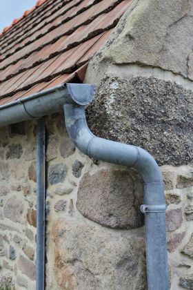 Mur en pierre avec gouttière et descente pluviale grises sous un toit en tuiles