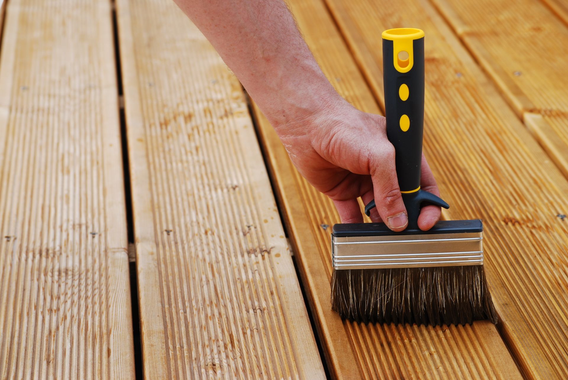 Application manuelle de teinture au pinceau sur une terrasse en bois, avec des planches neuves et non finies côte à côte.