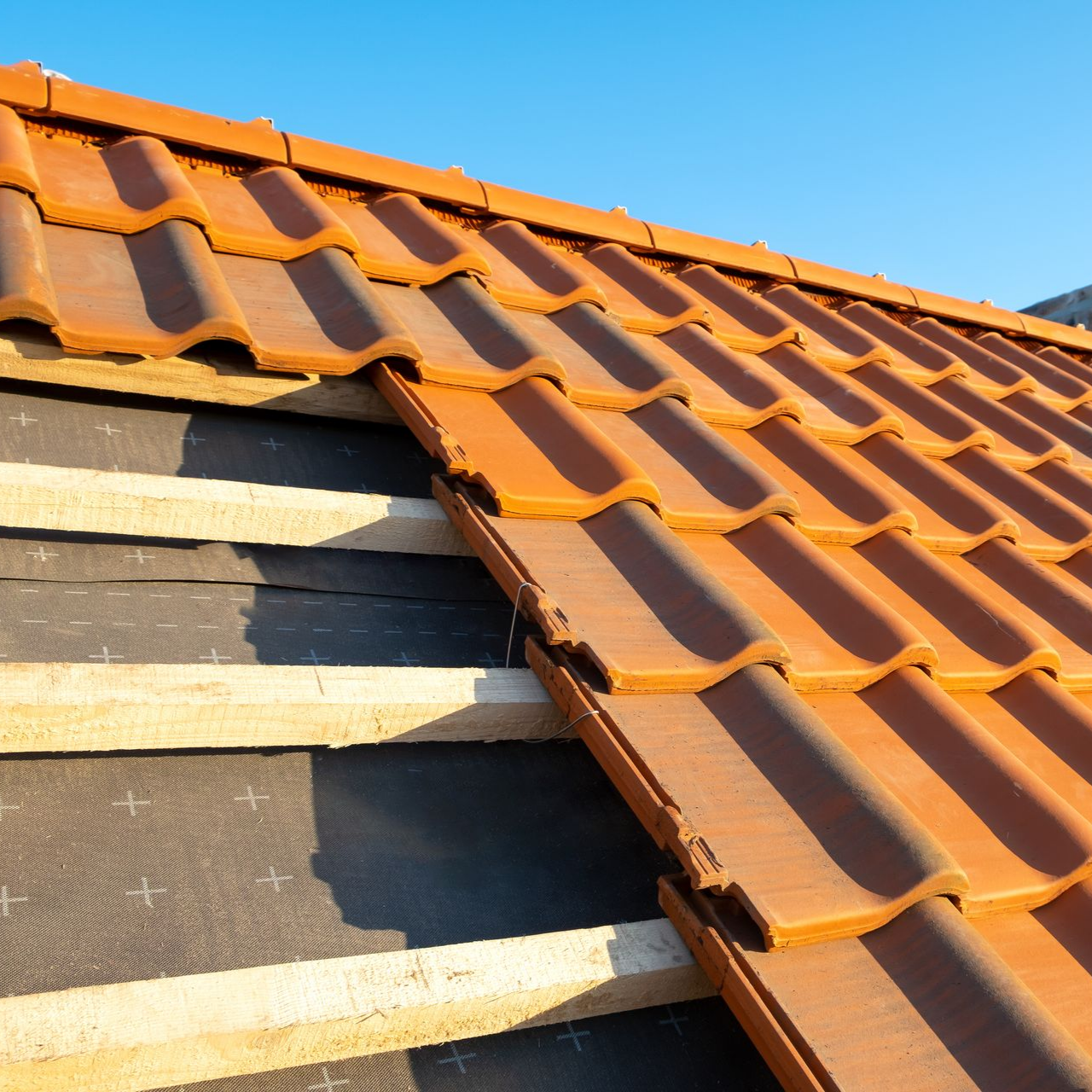 Des tuiles orange sont installées sur le toit d'une maison partiellement couverte, sous un ciel bleu.