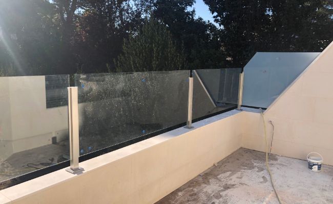 Un balcón con barandilla de cristal y árboles al fondo.