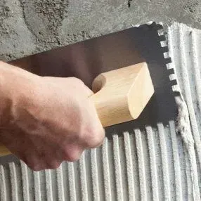 Una persona está usando una paleta para esparcir adhesivo para baldosas en un piso de baldosas.