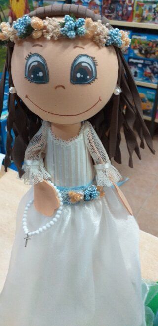 Muñeca grande con corona de flores, vestido de cuentas y bata blanca en un colorido escaparate.