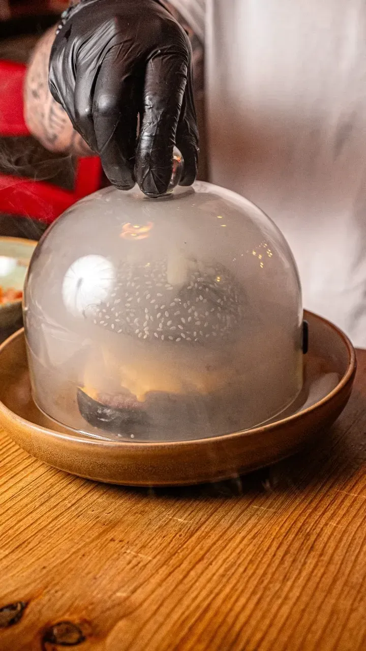 Una persona sostiene una hamburguesa debajo de una cúpula de plástico sobre un plato de madera.