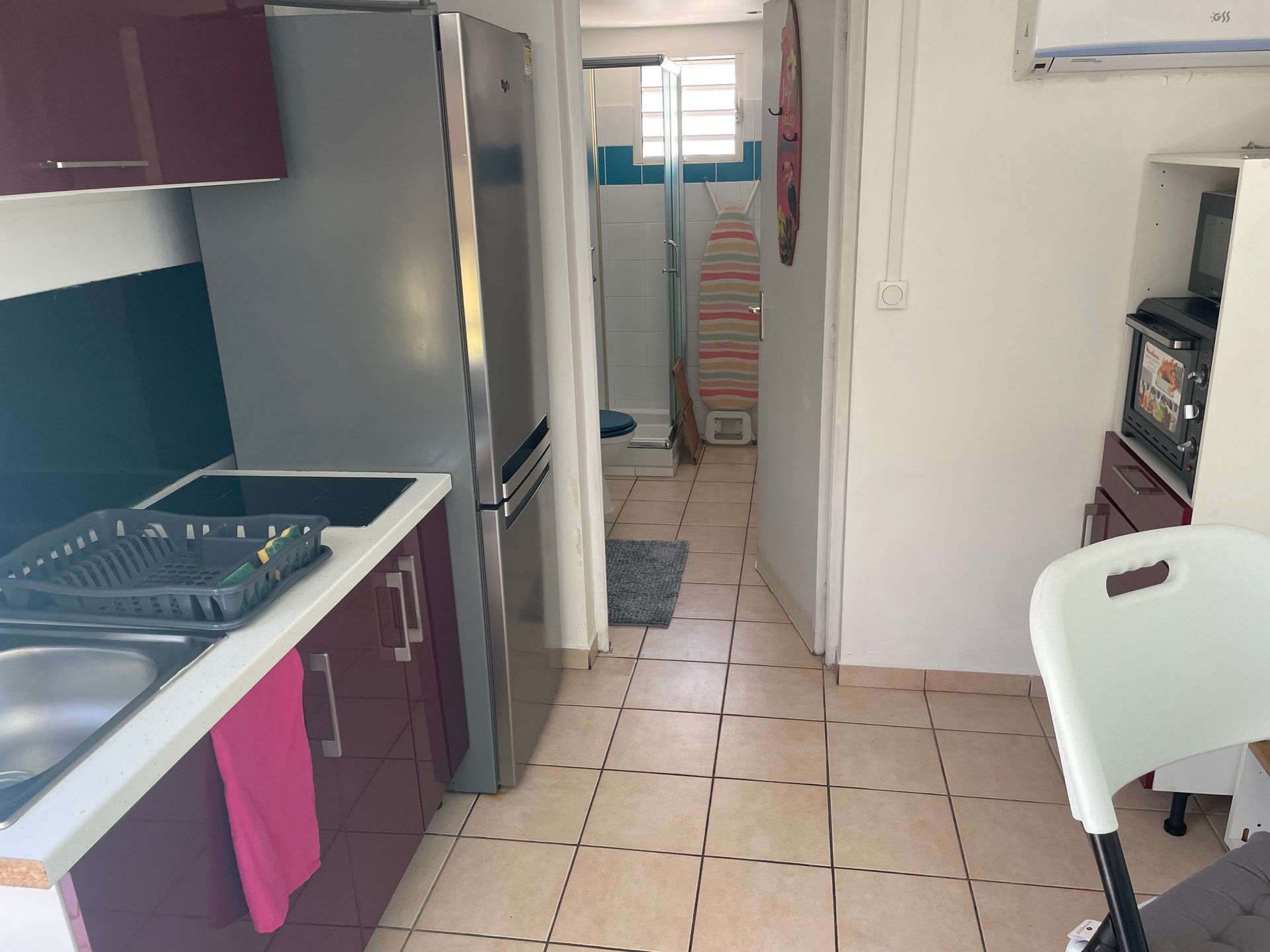 Cuisine étroite avec sol carrelé, évier et placards à gauche, réfrigérateur au centre et chaise près du four à droite.