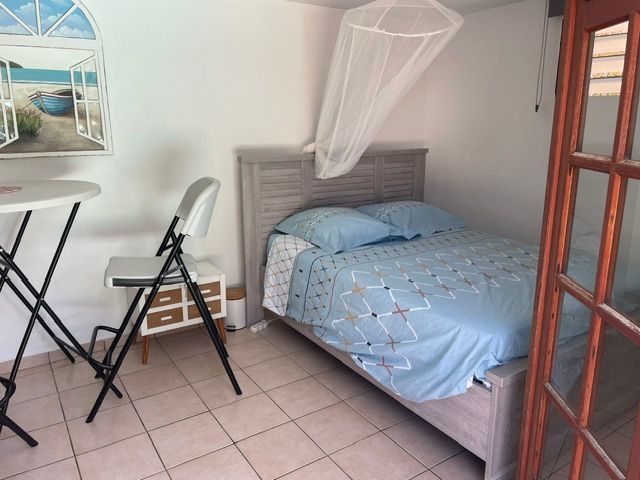 Petite chambre avec un lit simple, draps bleus, chaise pliante, table et moustiquaire à la fenêtre.