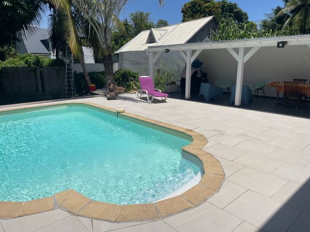 Piscine dans le jardin avec terrasse, pergola blanche et chaise longue rose baignée de soleil.