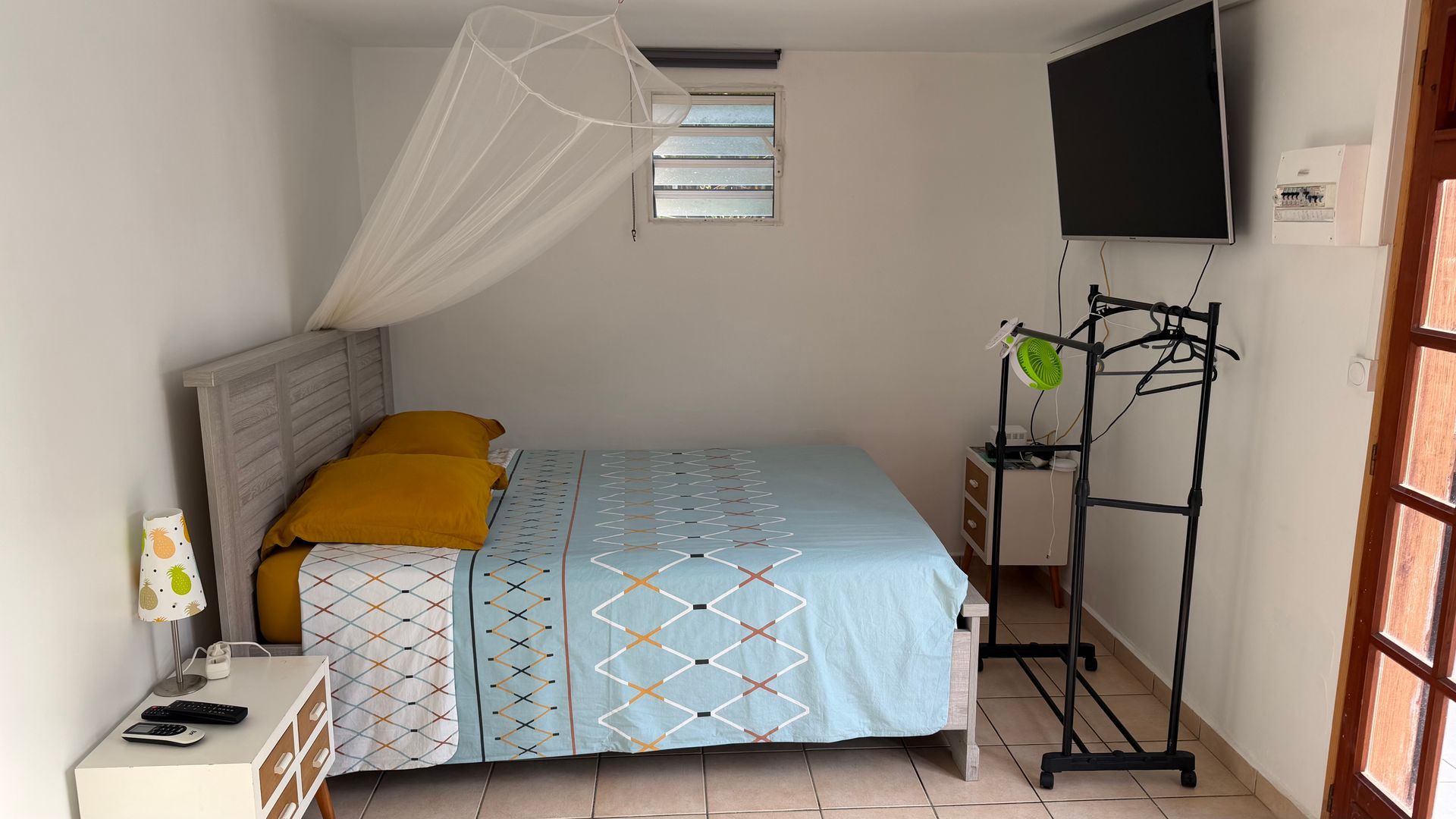 Petite chambre avec lit, moustiquaire, télévision murale et étagère près d'une fenêtre.