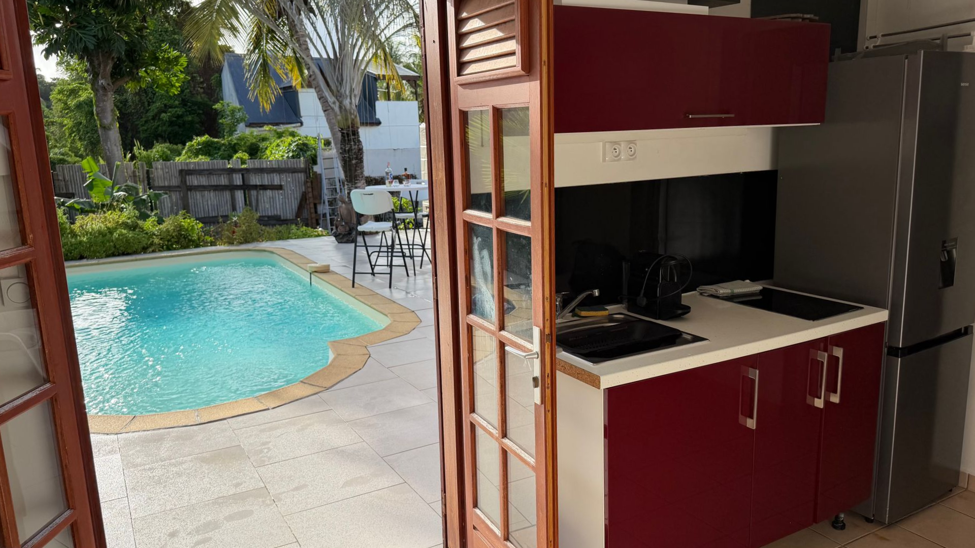 Vue sur la piscine à travers les portes ouvertes donnant sur une kitchenette rouge compacte et un réfrigérateur en acier inoxydable