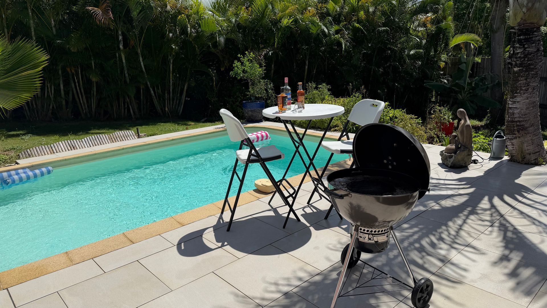Terrasse arrière avec une table au bord de la piscine, deux chaises et un barbecue au charbon de bois baigné de soleil