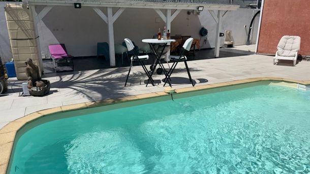 Piscine dans le jardin avec terrasse ombragée, chaises longues et petite table sous une pergola.