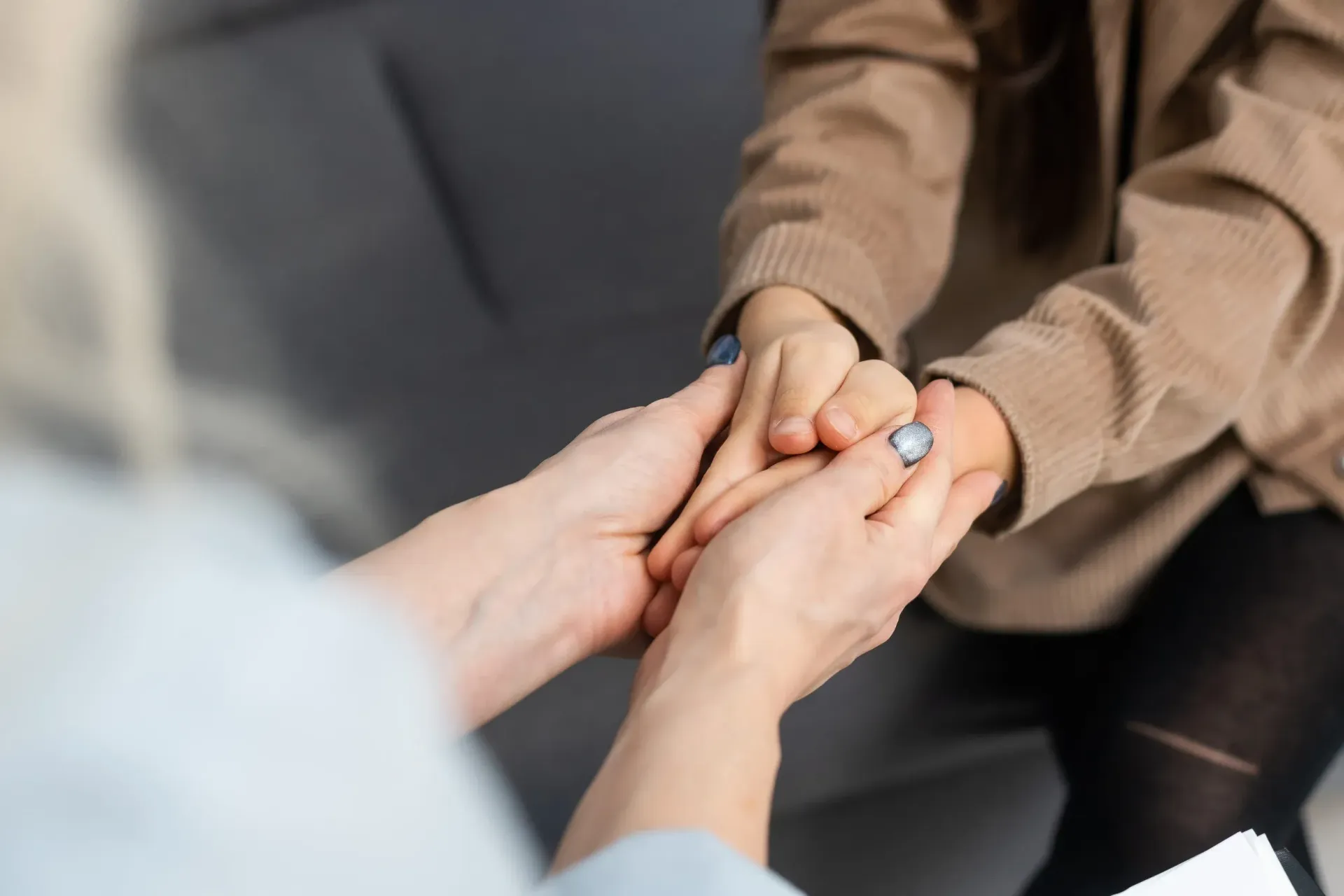 Dos pares de manos unidas en un gesto de consuelo y apoyo, sobre un fondo gris claro.