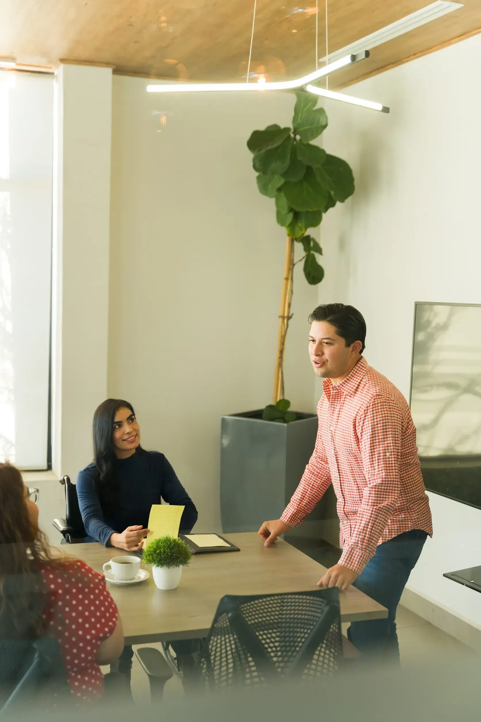 Tres compañeros colaboran alrededor de una mesa de oficina moderna con una gran planta en maceta cerca.