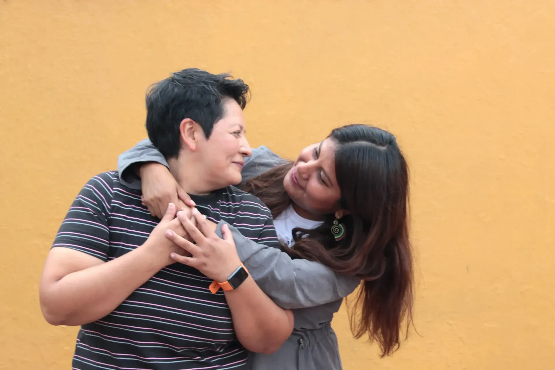 Dos personas se abrazan contra una pared amarilla lisa, sonriéndose mutuamente.