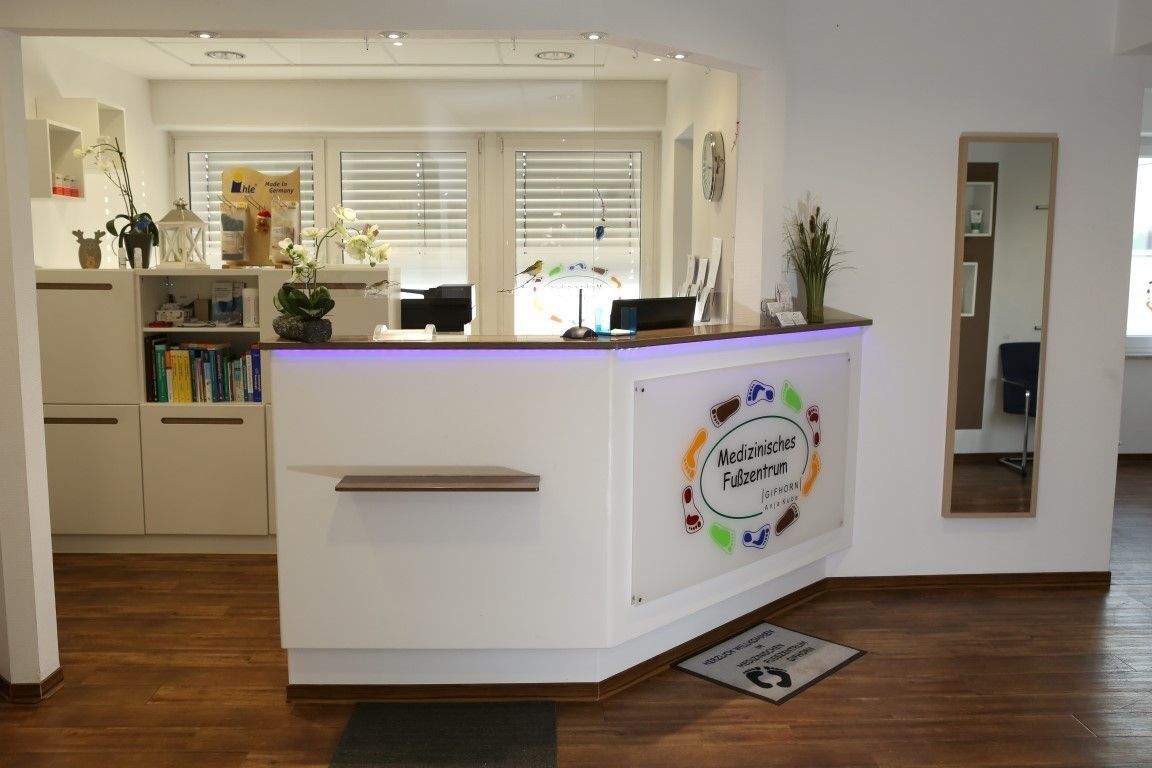 Empfangsbereich: Weißer Schreibtisch mit Logo, Regale, Spiegel, Holzboden und Sonnenlicht durch das Fenster.