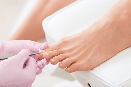 Fußpflegerin bearbeitet mit einem Werkzeug den Zehennagel einer Kundin, Nahaufnahme mit rosa Handschuhen und einem weißen Pedikürestuhl.