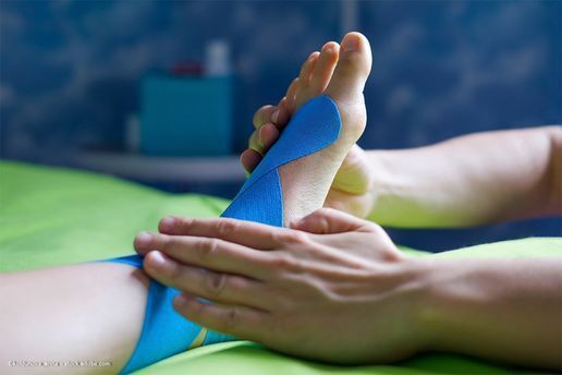 Der Fuß einer Person wird von zwei Händen auf einer grünen Oberfläche mit blauem Kinesiologie-Tape befestigt.