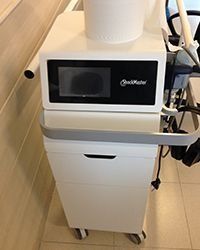 Dispositivo médico blanco sobre un carrito con ruedas en un pasillo, con pantalla negra y accesorios laterales.
