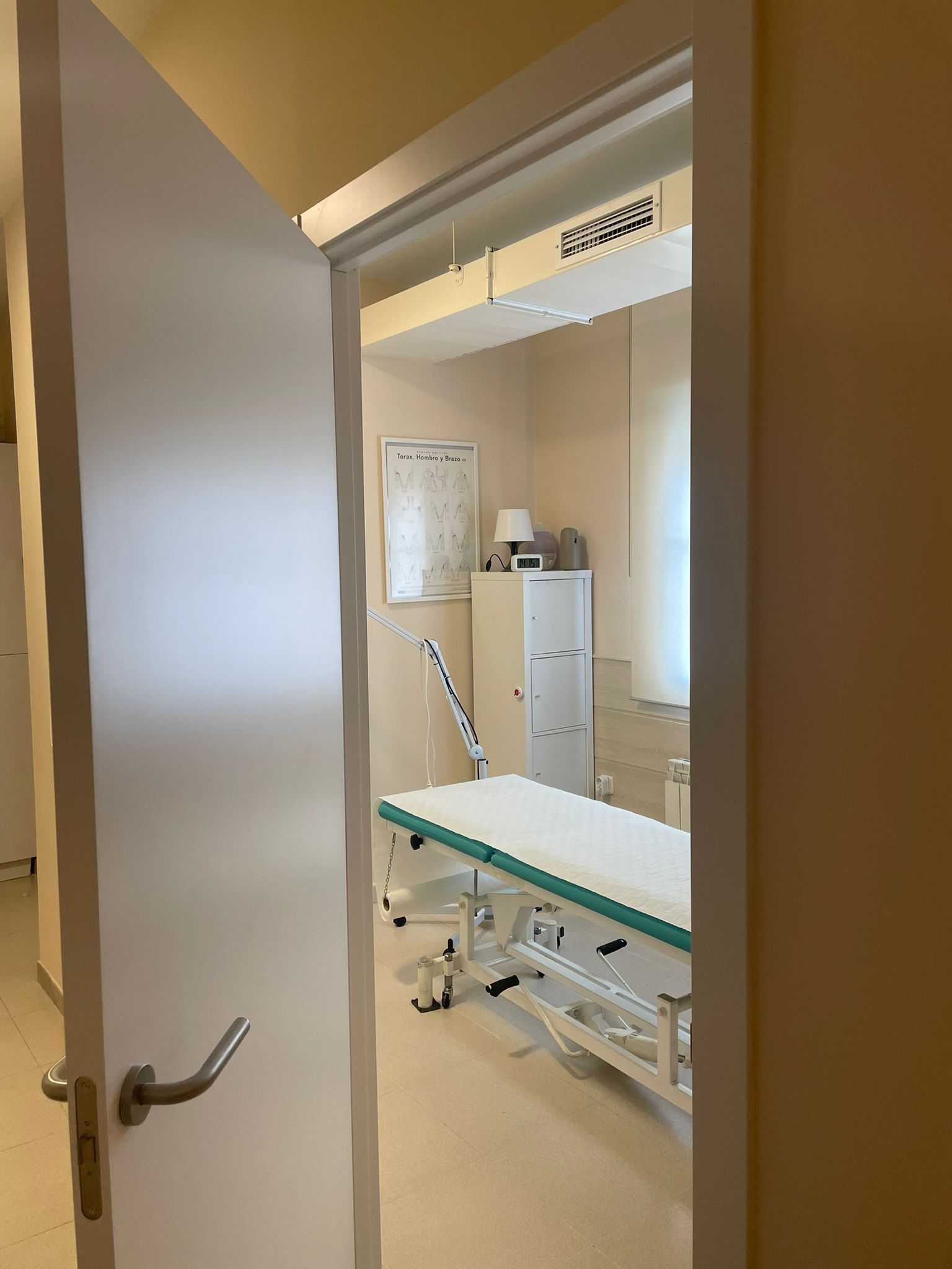 Sala de examen médico vacía con camilla, armario y pizarra blanca visibles a través de una puerta abierta.