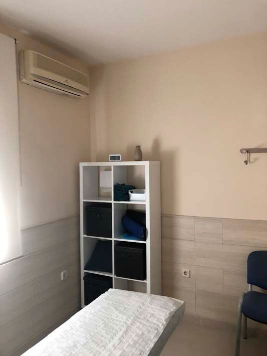 Habitación pequeña con una cama, una estantería blanca, aire acondicionado de pared y una silla azul junto a una pared de azulejos.
