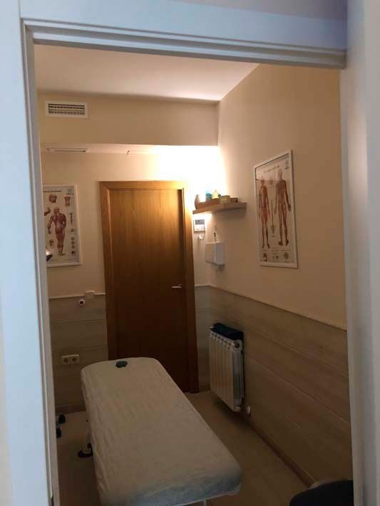 Sala de examen médico estrecha con camilla de tratamiento, puerta de madera cerrada, carteles en la pared y radiador bajo iluminación tenue.
