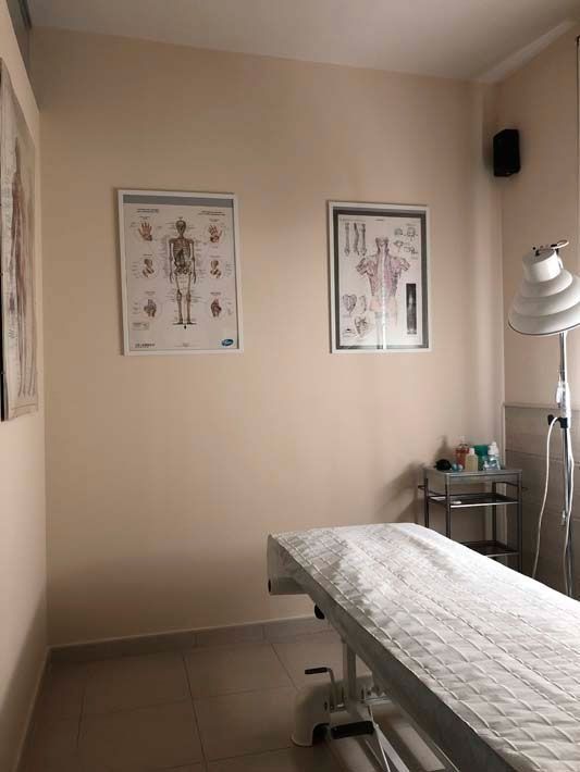 Sala de tratamiento con camilla de exploración, láminas anatómicas en la pared y lámpara médica.