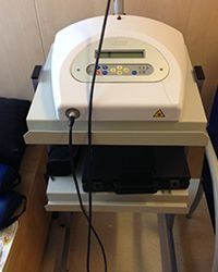 Dispositivo médico blanco sobre un carrito con una pequeña pantalla, botones y cables en una sala de consulta.